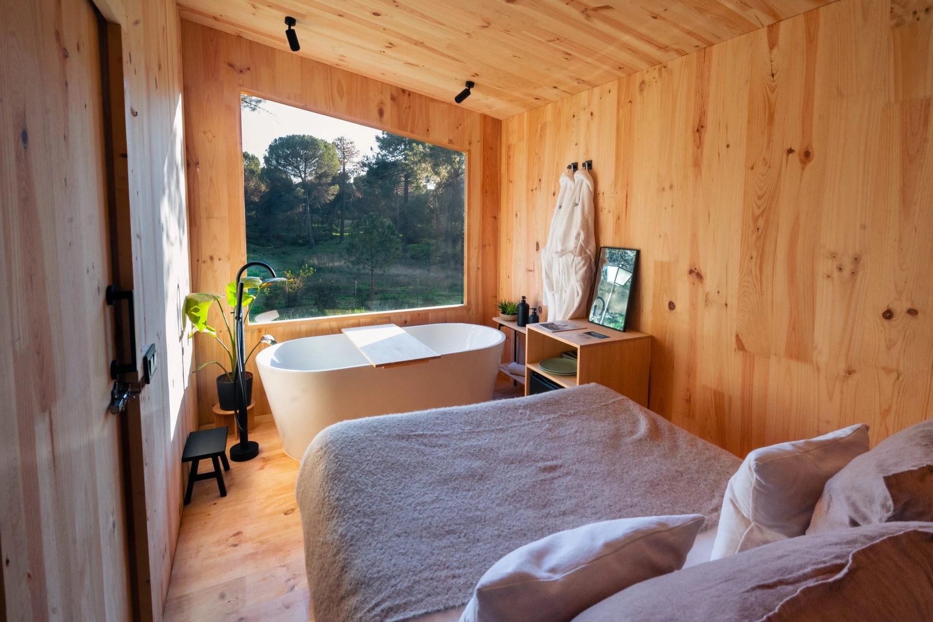 Interior de cabaña Kampaoh de madera con bañera exenta frente a un gran ventanal al bosque y cama de matrimonio